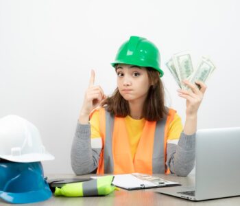 Female construction estimator in safety vest and green helmet holding dollar bills at desk with hard hats and laptop, symbolizing project cost planning.