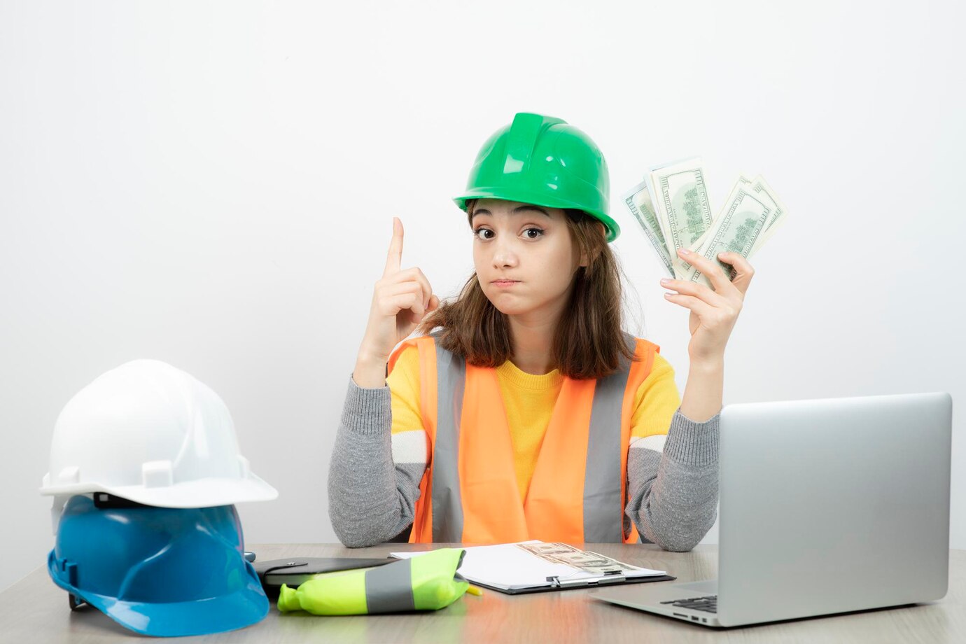 Female construction estimator in safety vest and green helmet holding dollar bills at desk with hard hats and laptop, symbolizing project cost planning.